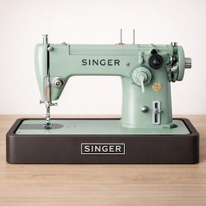 Könnte beinhalten: Eine mintgrüne Singer-Nähmaschine mit deutlich sichtbarem Markennamen. Die Maschine steht auf einem dunkelbraunen Sockel, der ebenfalls das Singer-Logo trägt. Sie verfügt über verschiedene Knöpfe und eine Nadel, was auf ihre Funktionalität hindeutet. SINGER 201 306 316 319 215r