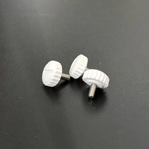 Könnte beinhalten: Drei kleine weiße Knöpfe mit einem kreisförmigen, geriffelten Rand und einer flachen Oberseite mit einem einzelnen horizontalen Schlitz. Jeder Knopf ist an einem kurzen, mit Gewinde versehenen Metallstift befestigt, der einen festen Halt bietet.