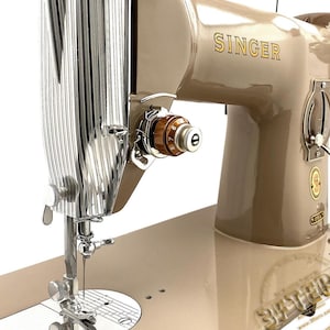 Könnte beinhalten: Eine Vintage Singer Nähmaschine in einem beigen Farbschema. Die Maschine verfügt über einen verchromten Nadelhalter und ein braunes und goldenes Spannrad. Das Wort "SINGER" ist in goldfarbenen Buchstaben auf dem Gehäuse der Maschine deutlich sichtbar. SINGER 201 306 316 319 215r