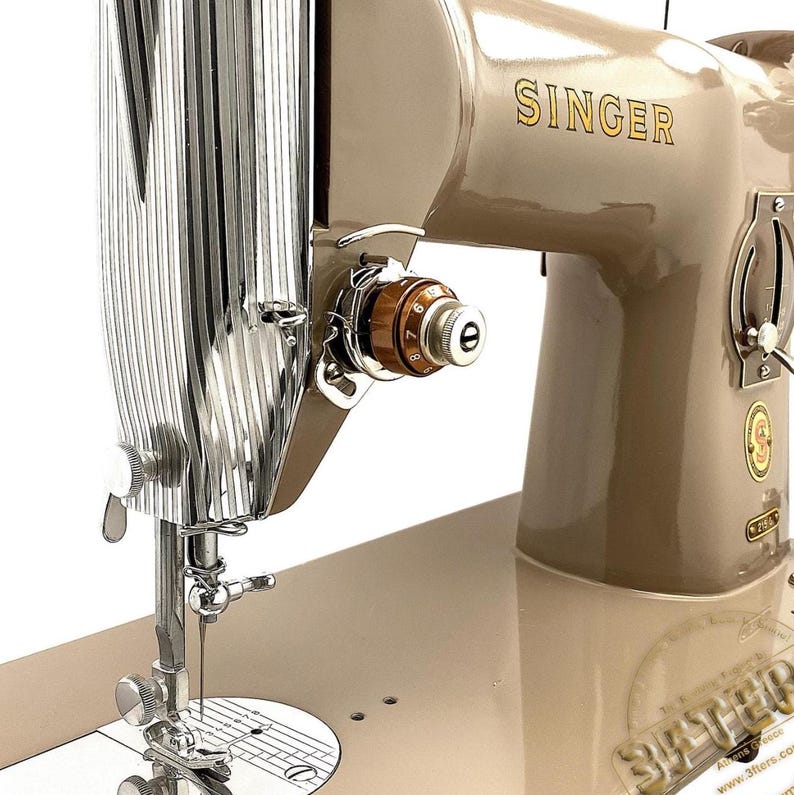 Könnte beinhalten: Eine Vintage Singer Nähmaschine in einem beigen Farbschema. Die Maschine verfügt über einen verchromten Nadelhalter und ein braunes und goldenes Spannrad. Das Wort "SINGER" ist in goldfarbenen Buchstaben auf dem Gehäuse der Maschine deutlich sichtbar. SINGER 201 306 316 319 215r