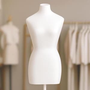 Op de afbeelding: Een witte paspop met een gladde afwerking. De vorm heeft een gedefinieerde buste, taille en heupen en is gemonteerd op een witte standaard. De achtergrond bevat een wazig beeld van andere paspoppen en kleding.