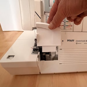 Puede incluir: Una remalladora Pfaff coverlock 4852 de color blanco con el panel frontal abierto. La máquina muestra el área de la aguja, el recorrido del hilo y una guía de puntadas impresa en la parte superior derecha. La carcasa es de plástico blanco.