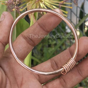 以下が含まれることがあります： ローズゴールドのバングルブレスレット、コイル状のアクセント付き。 ブレスレットは円形で、金属製に見えます。 ブレスレットには「TheJewelryCraftIN」の文字が見えます。 背景は緑の葉でぼやけています。