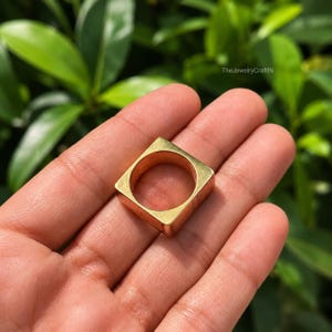 May include: A gold-toned ring featuring a square-shaped outer band with a circular interior opening. The surface of the metal is smooth and reflective, showcasing a geometric design. The text TheJewelryCraftIN is visible in the upper right corner.
