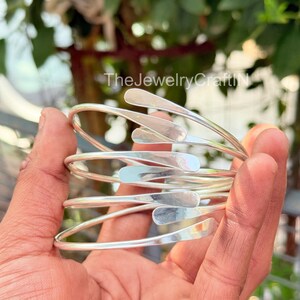 Könnte beinhalten: Eine Ansammlung silberfarbener Armreifen. Jeder Armreif hat ein gebogenes, offenes Design mit einem tropfenförmigen Akzent. Die Armreifen werden in einer Hand gehalten, mit einem verschwommenen grünen Hintergrund.