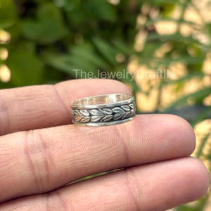 May include: A silver ring featuring a leaf design encircling the band. The ring is held between fingers, with a blurred green backdrop. The Jewelry CraftN is visible in the background.