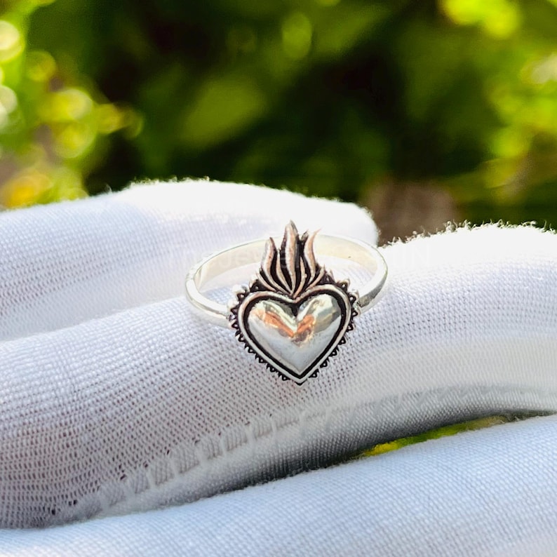 May include: A silver ring featuring a heart-shaped design with a flame motif at the top. The heart is detailed with a beaded border. The ring is shown on a white surface.