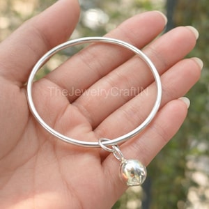 Può includere: Un bracciale rigido in argento con un piccolo ciondolo rotondo. Il bracciale ha una finitura liscia e lucida. Il ciondolo è di forma sferica, aggiungendo un dettaglio. Il logo The Jewelry Craft è visibile. Un gioiello semplice ed elegante.
