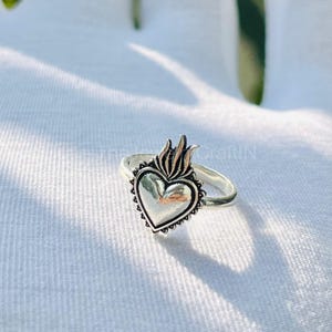 May include: A silver ring featuring a heart-shaped design with a flame detail at the top. The heart has a textured border and is set on a simple band. The ring is displayed on a white surface.