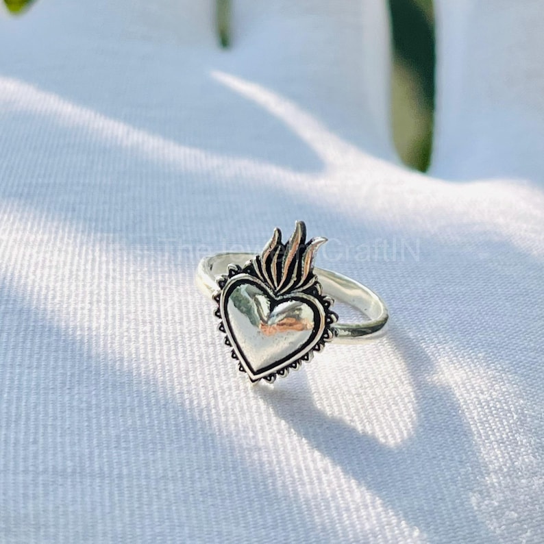 May include: A silver ring featuring a heart-shaped design with a flame detail at the top. The heart has a textured border and is set on a simple band. The ring is displayed on a white surface.