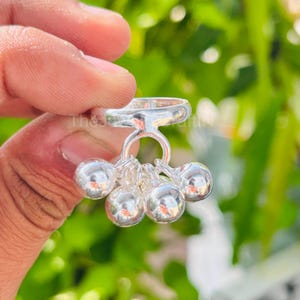 Può includere: Un anello d'argento con un gruppo di cinque piccole campane rotonde pendenti. L'anello è tenuto contro uno sfondo verde sfocato, suggerendo un ambiente esterno. Le campane sono lucide e riflettenti.