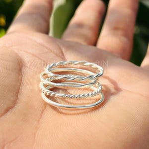 May include: A stack of four silver rings, each with a different design. Two rings have a twisted rope design, and the other two are smooth. The rings are displayed on a person's open palm.