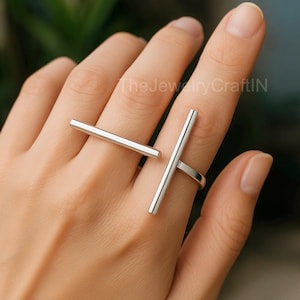 Puede incluir: Dos anillos de barra plateados, uno en un dedo y el otro al lado. Los anillos tienen un diseño minimalista, con una barra recta que se extiende por el dedo. The JewelryCraftIN es visible en el fondo.