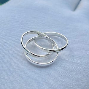 May include: A silver-colored ring with three interlocking bands. The bands are smooth and rounded, creating a continuous, intertwined design. The ring is displayed on a light blue fabric background.