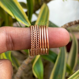 May include: A selection of copper-toned rings, including twisted rope designs, displayed on a finger. The rings have varying textures, with some smooth and others patterned. The background shows green and yellow leaves, and the text "The Jewelry Craft N".