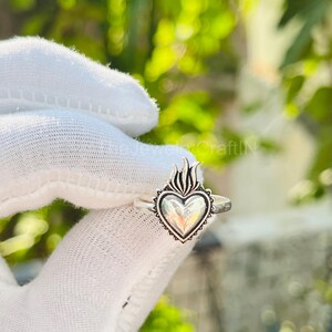 May include: A silver ring featuring a heart-shaped design with flames at the top. The heart has a detailed border and is set on a thin band. The ring is displayed against a white background.