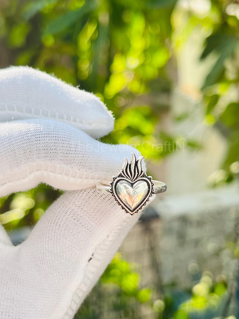 May include: A silver ring featuring a heart-shaped design with flames at the top. The heart has a detailed border and is set on a thin band. The ring is displayed against a white background.