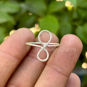 May include: A silver-colored ring with an infinity symbol design. The ring is thin and delicate, with a polished finish. The ring is held in a hand, with a blurred green background.