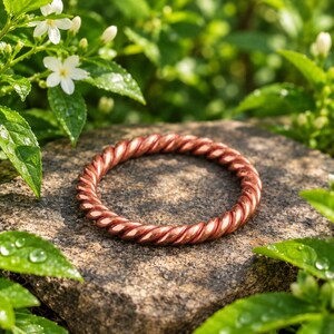 Pode incluir: Uma pulseira circular feita de fio de cobre trançado com um acabamento metálico castanho-avermelhado. A banda apresenta uma textura contínua semelhante a uma corda criada pelos fios de metal em espiral, formando um anel sólido e uniforme.