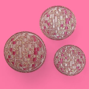 Può includere: Tre bottoni rotondi decorativi con un motivo intrecciato rosa e bianco e dettagli glitterati dorati. I bottoni sono disposti su uno sfondo rosa acceso. I bottoni variano di dimensioni.