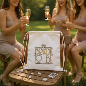 Może przedstawiać: Kremowa torba ze sznurkiem z napisem "BRIDE TO BE" w złotych literach i diamentową grafiką. Torba leży na drewnianym krześle, a w tle trzy kobiety trzymają kieliszki do szampana.