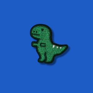 Könnte beinhalten: Ein grüner Dinosaurier-Aufnäher mit schwarzem Umriss. Der Dinosaurier hat einen freundlichen Ausdruck und weiße Akzente auf seinem Schwanz. Der Aufnäher ist vor einem einfarbigen blauen Hintergrund platziert.