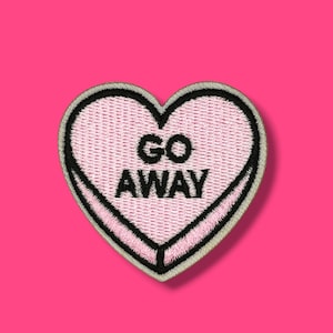 Könnte beinhalten: Ein rosa herzförmiger, gestickter Aufnäher mit den schwarzen Worten "GO AWAY". Der Aufnäher hat einen schwarzen Rand und einen rosa Hintergrund, vor einem hellrosa Hintergrund.