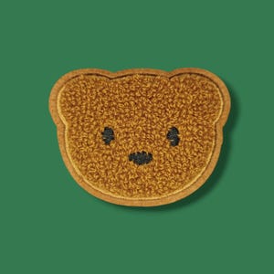 Könnte beinhalten: Ein brauner Teddybär-Aufnäher mit strukturiertem, plüschigem Aussehen. Der Bär hat schwarze Augen und eine schwarze Nase mit einer gestickten Kontur. Der Aufnäher ist vor einem einfarbigen grünen Hintergrund platziert.