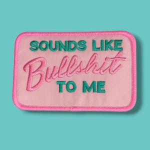 Può includere: Una toppa rettangolare ricamata con sfondo rosa chiaro e bordo cucito rosa scuro. Il testo recita "SOUNDS LIKE" in verde acqua, "Bullshit" in grande corsivo rosa e "TO ME" in verde acqua, tutto centrato sulla superficie del tessuto.