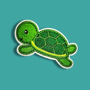 Può includere: Una toppa ricamata a forma di tartaruga con testa e arti verde brillante, un guscio verde scuro con motivo esagonale e una sottile striscia gialla sul lato, il tutto delineato da un bordo bianco spesso e cuciture nere.