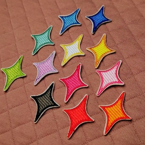 Könnte beinhalten: Eine Ansammlung von gestickten sternförmigen Aufnähern in verschiedenen Farben wie Blau, Grün, Rosa, Gelb, Rot und Orange. Jeder Aufnäher hat einen weißen Rand und liegt auf einem gesteppten, braunen Stoff. Ideal zum Verzieren von Kleidung oder Accessoires.