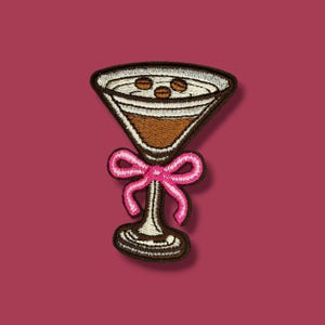 Può includere: Toppa ricamata raffigurante un bicchiere da martini marrone con un fiocco rosa. Il bicchiere contiene un liquido marrone e tre chicchi di caffè. La toppa ha un bordo marrone scuro su sfondo bordeaux.