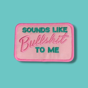Może przedstawiać: Haftowana naszywka z różowym tłem i różową obwódką. Naszywka zawiera napis "SOUNDS LIKE Bullshit TO ME" w kolorze turkusowym i różowym. Naszywka jest prostokątna i ma turkusowe tło.