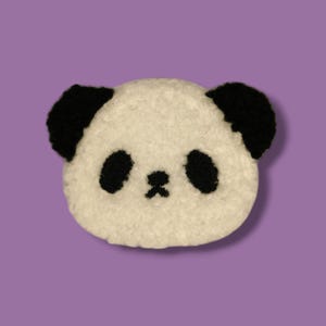 Könnte beinhalten: Ein rundes, weißes und schwarzes Pandagesicht mit schwarzen Ohren, Augen und Nase. Das Gesicht des Pandas besteht aus einem strukturierten, flauschigen Material. Der Hintergrund ist einfarbig lila.