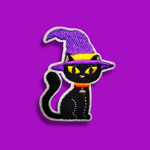 Könnte beinhalten: Ein gestickter Aufnäher, der eine schwarze Katze mit einem violetten Hexenhut mit gelbem Band zeigt. Die Katze hat leuchtend gelbe Augen, ein orangefarbenes Halsband mit einem kleinen Anhänger und einen geschwungenen Schwanz, alles weiß umrandet.