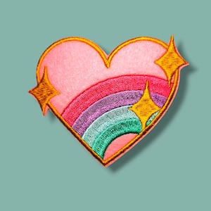 Può includere: Una toppa ricamata a forma di cuore con una sezione superiore rosa chiaro e un arcobaleno multicolore nelle tonalità del rosso, viola e verde acqua. Il design è delineato con filo dorato e accentuato da due stelle dorate ricamate a quattro punte.
