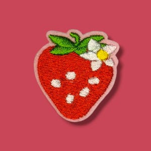Op de afbeelding: Een geborduurde patch in de vorm van een rode aardbei met groene blaadjes en een witte bloem met een geel hartje. De aardbei heeft witte geborduurde stippen die zaden voorstellen en is aan alle kanten omrand met een lichtroze rand.