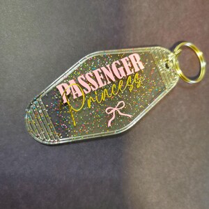 Könnte beinhalten: Transparenter Acryl-Schlüsselanhänger mit den Worten "PASSENGER Princess" in rosa und goldener Schrift, akzentuiert mit einer rosa Schleife. Der Schlüsselanhänger ist mit bunten Glitzer gefüllt und hat einen goldenen Schlüsselring.