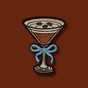 Könnte beinhalten: Gestickter Aufnäher mit einem Cocktail in einem Martiniglas mit drei Kaffeebohnen. Das Glas ist braun mit einem weißen Rand und einer hellblauen Schleife. Der Aufnäher hat einen schwarzen Rand und ist vor einem braunen Hintergrund platziert.