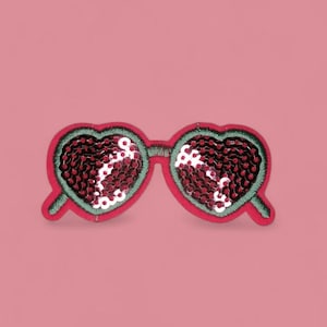 Könnte beinhalten: Ein herzförmiger Pailletten-Aufnäher, der einer Sonnenbrille ähnelt. Der Aufnäher hat einen roten Umriss mit grünen Nähten und ist mit roten und weißen Pailletten gefüllt. Der Hintergrund ist einfarbig rosa.