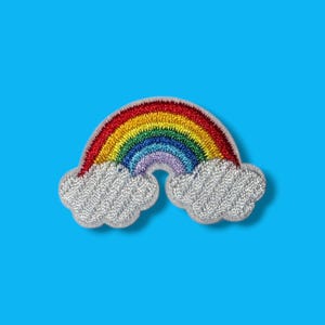Puede incluir: Un parche bordado que presenta un arcoíris colorido arqueado entre dos nubes blancas. El arcoíris consta de hilos rojos, naranjas, amarillos, verdes, azules y morados. Las nubes están texturizadas con hilo blanco plateado y tienen un borde festoneado.