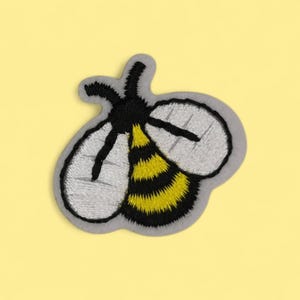 Könnte beinhalten: Gestickter Bienenaufnäher mit grauem Umriss, schwarzem Kopf und Streifen, gelb-schwarz gestreiftem Hinterleib und grauen Flügeln. Der Aufnäher befindet sich auf einem gelben Hintergrund. Ideal für Kleidung oder Accessoires.