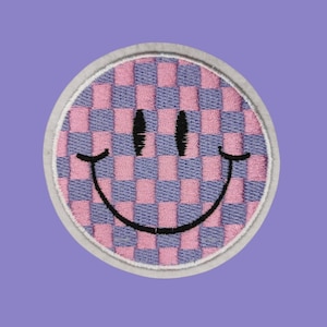 Könnte beinhalten: Gestickter Aufnäher mit einem Smiley-Gesicht-Design. Das Gesicht hat ein rosa-blaues Schachbrettmuster, schwarze Augen und ein Lächeln. Der runde Aufnäher hat einen weißen Rand, vor einem lila Hintergrund.