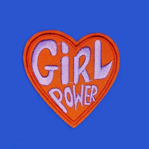 Puede incluir: Un parche bordado en forma de corazón, con las palabras "GIRL POWER" en letras moradas sobre un fondo naranja. El parche está sobre un fondo azul liso.