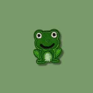 Könnte beinhalten: Gestickter Aufnäher mit einem lächelnden grünen Frosch mit weißen Augen und einem hellgrünen Bauch. Der Froschaufnäher befindet sich auf einem einfarbigen grünen Hintergrund. Ideal zum Aufnähen auf Kleidung, Taschen oder andere Stoffartikel.