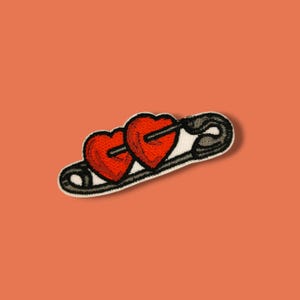 Könnte beinhalten: Gestickter Aufnäher mit zwei roten Herzen, die von einer schwarzen Sicherheitsnadel durchbohrt werden. Die Herzen haben eine strukturierte Optik und sind schwarz umrandet. Die Sicherheitsnadel ist schwarz und weiß, vor orangefarbenem Hintergrund.