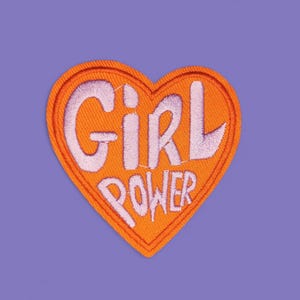 Könnte beinhalten: Ein orangefarbener herzförmiger Aufnäher mit den Worten "GIRL POWER" in Weiß gestickt. Der Aufnäher befindet sich auf einem einfarbigen lila Hintergrund. Dieser Aufnäher ist für Kleidung oder Accessoires geeignet.
