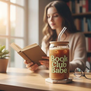 Op de afbeelding: Een doorzichtig glas met een bamboe deksel en een glazen rietje, gevuld met ijskoffie. Op de voorkant van het glas staat de groene tekst "Book Club Babe". Het glas staat op een witte zeshoekige onderzetter op een houten oppervlak.