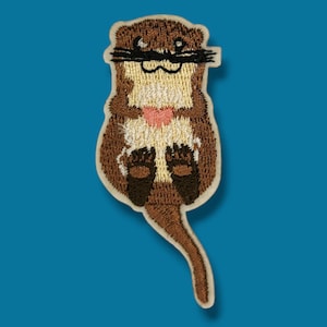 Könnte beinhalten: Gestickter Aufnäher mit einem Cartoon-Otter, der ein rosa Herz hält. Der Otter hat braunes Fell, einen weißen Bauch und schwarze Schnurrhaare. Der Aufnäher hat einen cremefarbenen Rand und ist vor einem blauen Hintergrund platziert.