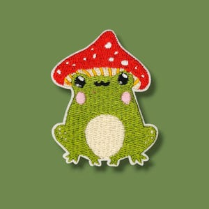 Può includere: Toppa ricamata raffigurante una rana verde con un cappello a fungo rosso a pois bianchi. La rana ha la pancia color crema, guance rosa e occhi neri. La toppa è bordata di bianco su sfondo verde.